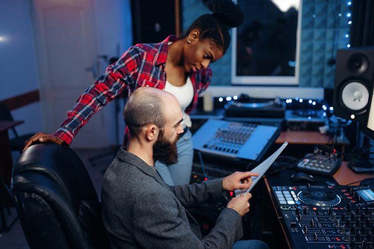 A broadcasting producer and singer working together on a script in the studio, surrounded by mixers and audio equipment for an audio engineering degree.